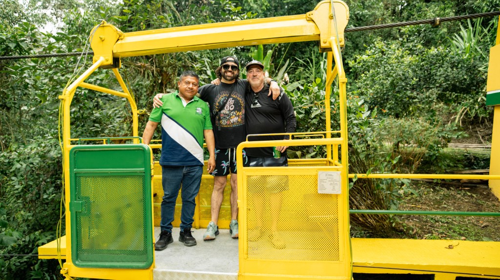 Three men pose together in a yellow gondola surrounded by lush green foliage.